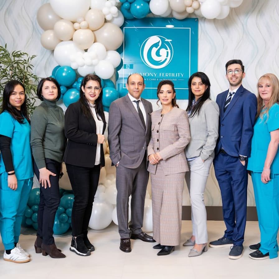 Nine people standing indoors in front of a blue sign reading "Harmony Fertility" with white, beige, and teal balloons arranged around it.
